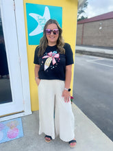 Load image into Gallery viewer, Woman wearing a black t-shirt with a colorful design and white pants standing in front of a yellow wall with a blue sign.