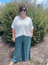 Load image into Gallery viewer, woman wearing a ivory top and jade pants standing outdoors with greenery in the background
