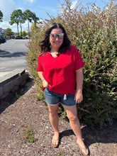 Load image into Gallery viewer, woman wearing a red shirt and denim shorts standing outdoors with trees in the background
