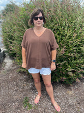 Load image into Gallery viewer, Woman wearing a brown top and white shorts standing in front of a bush.