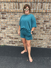 Load image into Gallery viewer, Woman wearing a teal outfit standing against a brick wall.