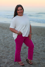 Load image into Gallery viewer, Woman standing on a beach wearing a white top and pink pants with a sunset in the background.