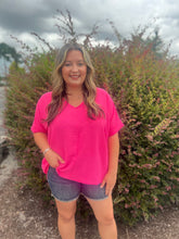 Load image into Gallery viewer, Woman wearing a pink shirt and denim shorts standing in front of a bush with green and purple leaves.