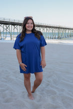 Load image into Gallery viewer, Woman in a blue dress standing on a sandy beach with a pier in the background