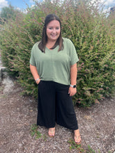 Load image into Gallery viewer, Woman wearing a green top and black pants standing in front of bushes.