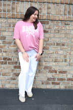 Load image into Gallery viewer, Woman standing against a brick wall wearing a pink amen graphic shirt tee and white pants.