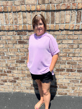 Load image into Gallery viewer, Person wearing a light purple shirt and black shorts standing against a brick wall.