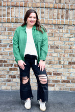 Load image into Gallery viewer, Woman wearing a green long sleeve gauze button up top , white shirt, and black ripped jeans standing against a brick wall.