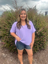 Load image into Gallery viewer, Woman wearing a purple shirt and denim shorts standing in front of bushes with a cloudy sky.