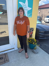Load image into Gallery viewer, Woman standing outside a store with a sign in the background wearing a thankful rust tshirt with black pants