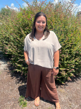 Load image into Gallery viewer, Woman wearing a beige top and brown pants standing in front of green bushes.