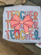 Load image into Gallery viewer, Light Blue Stacked Teacher Tee laying on a couch