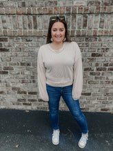 Load image into Gallery viewer, Woman wearing a beige sweater and blue jeans standing against a brick wall.
