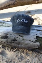 Load image into Gallery viewer, Black cap with white text on a piece of driftwood
