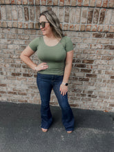 Load image into Gallery viewer, Woman wearing a green top and blue jeans standing against a brick wall.
