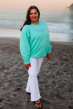 Load image into Gallery viewer, Woman wearing a teal sweatshirt and white pants standing on a beach.
